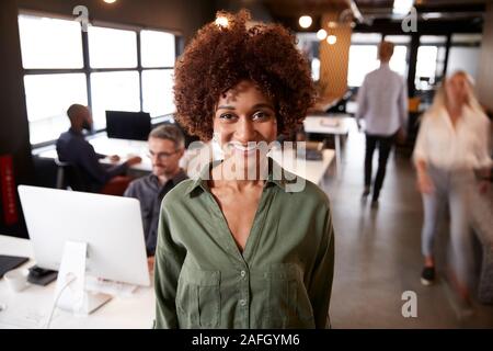 La millenaria femmina nero creative in piedi in un affollato ufficio casual, sorridente alla fotocamera