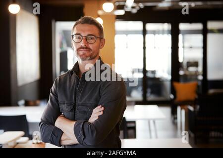 Di mezza età maschio bianco creative con gli occhiali in piedi in un ufficio cercando di fotocamera, vita fino Foto Stock