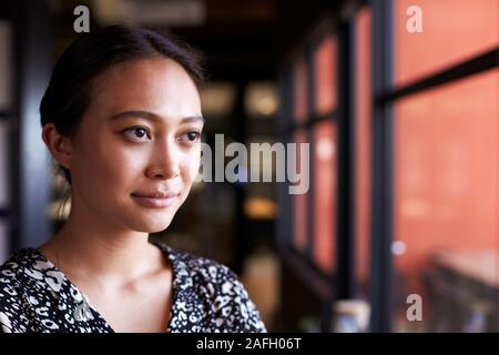 La millenaria Asian imprenditrice guardando fuori della finestra in un ufficio, testa e spalle, close up Foto Stock