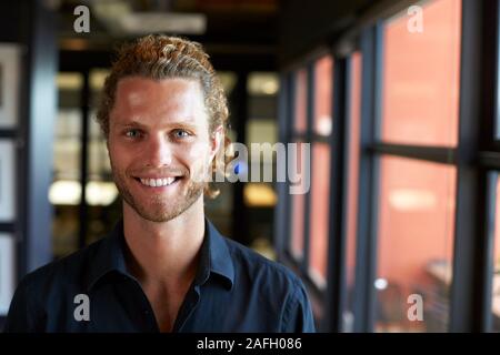 La millenaria maschio bianco creative business sorridere alla telecamera tramite la finestra in un ufficio, close up Foto Stock