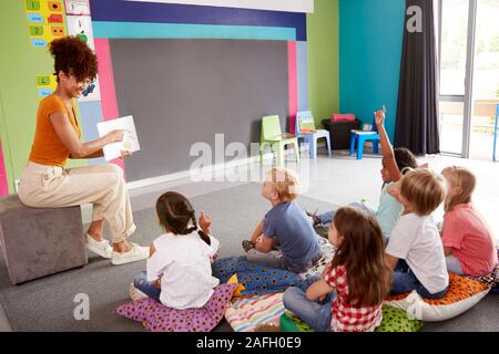 Gli alunni elementari alzando le mani per rispondere alla domanda più femminile insegnante legge la storia in aula Foto Stock