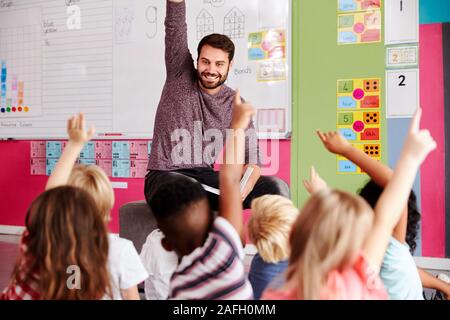 Gli alunni elementari alzando le mani per rispondere alla domanda come maschio insegnante legge la storia in aula Foto Stock