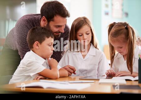 Insegnante di sesso maschile con tre scuole elementari gli alunni indossano uniformi utilizzando tavoletta digitale alla scrivania Foto Stock