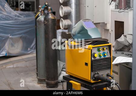 Un giallo macchina di saldatura con una maschera protettiva sulla sommità sorge in un workshop di metallo nei pressi di bombole di anidride carbonica. Industria e produzione. Costruzione Foto Stock