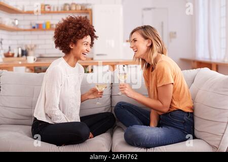 Due amiche rilassante sul divano di casa con un bicchiere di vino a parlare insieme Foto Stock