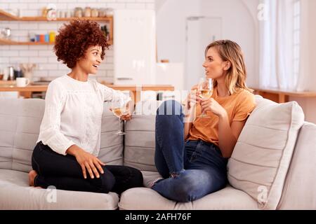 Due amiche rilassante sul divano di casa con un bicchiere di vino a parlare insieme Foto Stock