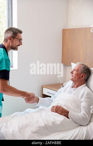 Chirurgo visitando e stringono le mani con i Senior paziente di sesso maschile nel letto di ospedale in unità Geriatrica Foto Stock