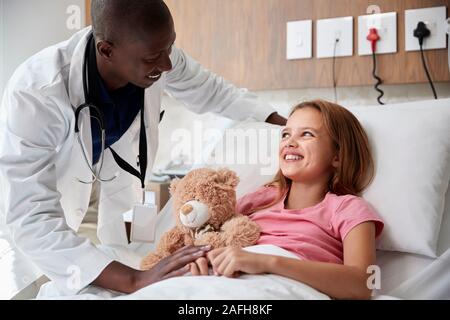 Medico maschio visitando ragazza distesa nel letto di ospedale abbracciando Teddy Bear Foto Stock