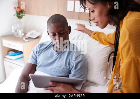 Medico con tavoletta digitale visitando e parlando con il paziente di sesso maschile nel letto di ospedale Foto Stock