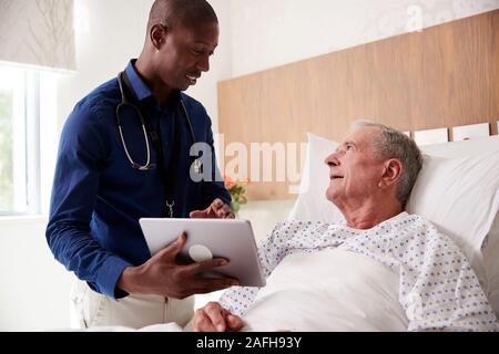 Medico con tavoletta digitale visitando e parlando con i Senior paziente di sesso maschile nel letto di ospedale Foto Stock