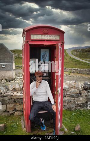 Uomo seduto in remoto tradizionale telefono rosso scatola avendo conversazione, Isle of Harris, Ebridi Esterni, Scozia Foto Stock