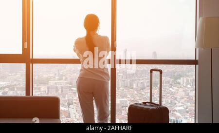 Bruna giovane donna viaggiatore con valigia in una camera di hotel con vista panoramica sulla citta'. Lei è in piedi vicino alla finestra e guardando alla città, alla luce del sole Foto Stock