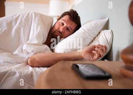 Metà uomo adulto dorme nel letto, guardando lo smartphone sul comodino in primo piano Foto Stock