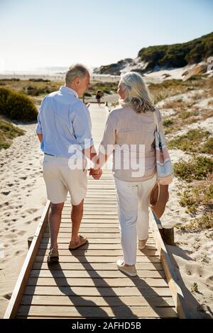 Senior bianco giovane camminare lungo la passeggiata a mare di legno su una spiaggia tenendo le mani a piena lunghezza e vista posteriore Foto Stock