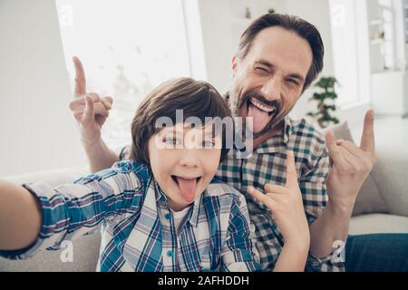 Hey There seguaci. Close up foto di positivo allegro bello ragazzo papà rendendo difficile simbolo punk e il suo piccolo figlio tenendo selfie sullo smartphone Foto Stock