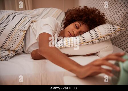 La millenaria americano africano donna mezzo addormentato nel letto, raggiungere fuori per la sveglia, close up Foto Stock