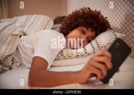 La millenaria americano africano donna mezzo addormentato nel letto, guardando il suo smartphone, close up Foto Stock
