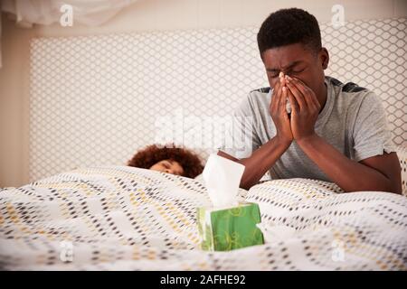 La millenaria African American uomo seduto a letto soffia il naso, mentre il suo partner dorme, close up Foto Stock