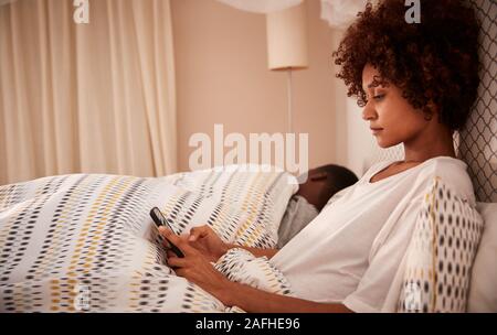 La millenaria americano africano donna seduta sul letto utilizzando il suo smartphone, vista laterale, vicino fino Foto Stock
