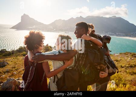 La millenaria amici su una escursione di abbracciare al vertice per celebrare la loro salita, vista posteriore Foto Stock