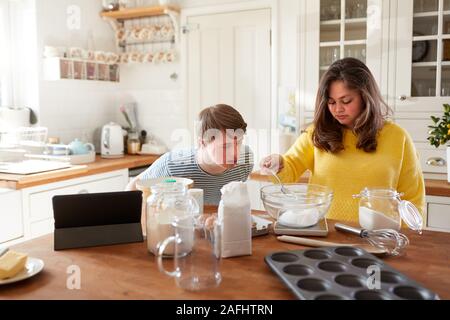 Giovani sindrome Downs giovane ricetta seguente sulla tavoletta digitale per cuocere la torta nella cucina di casa Foto Stock