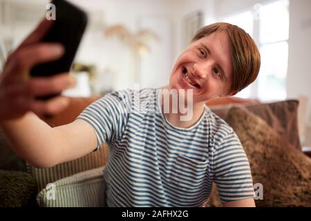 Giovani la sindrome di Down uomo seduto sul divano utilizzando il telefono cellulare per prendere Selfie a casa Foto Stock