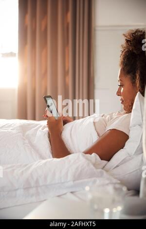 Donna seduta sul letto guardando il telefono cellulare dopo aver svegliato Foto Stock