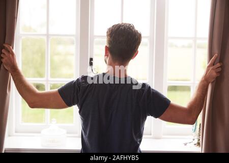 Vista posteriore dell'uomo indossando pigiami apertura camera da letto tende all inizio di un nuovo giorno Foto Stock