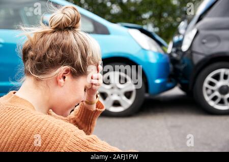 Automobilista femmina con testa in mani seduto accanto ai veicoli coinvolti in un incidente stradale Foto Stock