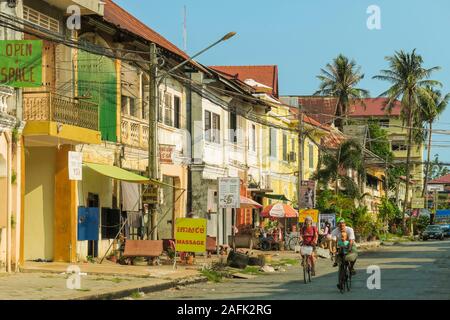 Genitori europei e bambini in bicicletta oltre il negozio fronti sulla strada 710 in questo vecchio porto coloniale francese fiume; Kambot, Provincia Kambot, Cambogia Foto Stock