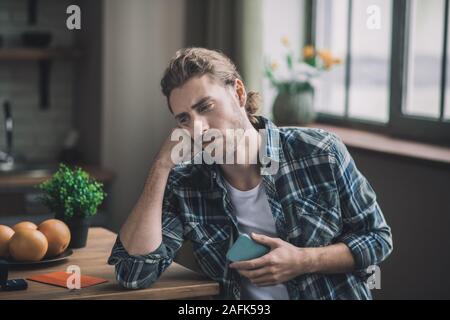 Annoiato giovane uomo seduto nella sua cucina con il suo smartphone Foto Stock