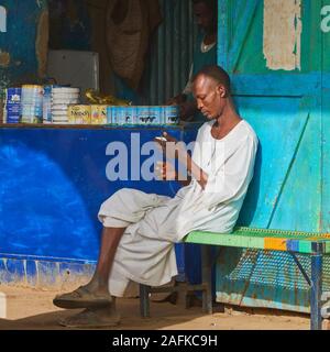 Il governo di Khartoum, Sudan, ca. Febbraio 8, 2019: uomo sudanese in abiti tradizionali si siede su un banco di lavoro nella parte anteriore del negozio per il latte in polvere e ascolta il suo mo Foto Stock