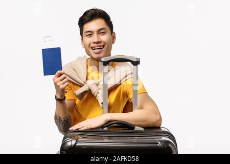 Attraente allegro giovani asiatici uomo seduto vicino al nero valigia, bagagli imballati, tenendo il passaporto e biglietto aereo, in attesa in aeroporto per il terminale Foto Stock