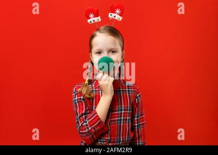 Funny felice bambina rendendo naso da palla di Natale. Un bambino in un abito rosso e manopole con archetto su uno sfondo colorato. Foto Stock