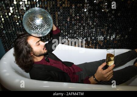 Stanco uomo con palla da discoteca dalla testa e flûte di champagne relax nella vasca da bagno Foto Stock