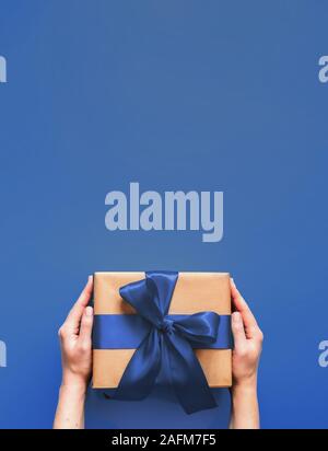 Mani femminili tenere confezione regalo su fondo blu intenso con copia spazio per il design. Ragazza caucasica mani confezione regalo nel mestiere della carta da imballaggio con la moda di colore blu 2020 nastro di raso. Verticale. Foto Stock