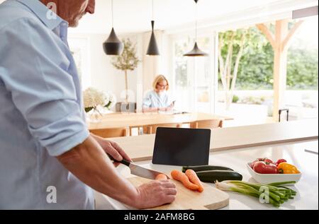 Senior uomo ricetta seguente sulla tavoletta digitale come moglie si siede al tavolo da pranzo una legge Magazine Foto Stock