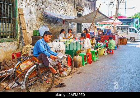 YANGON, MYANMAR - 17 febbraio 2018: il rickshaw attende i clienti leggendo il giornale su un angolo della squallida strada del mercato di Chinatown, su Foto Stock