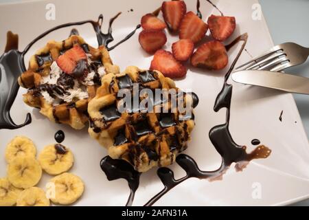 Cialde con lo sciroppo al cioccolato, crema, fragole e banane su una piastra Foto Stock