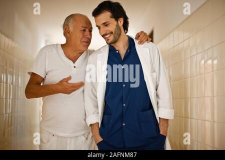 Maschio maturo paziente camminare e parlare con connettore maschio di tipo medico ospedale giù il corridoio. Foto Stock