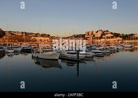 Yacht e altre imbarcazioni ormeggiate nel porto di Torquay, Devon, Regno Unito, mentre il sole tramonta illuminando la città dietro con luce arancione. Foto Stock