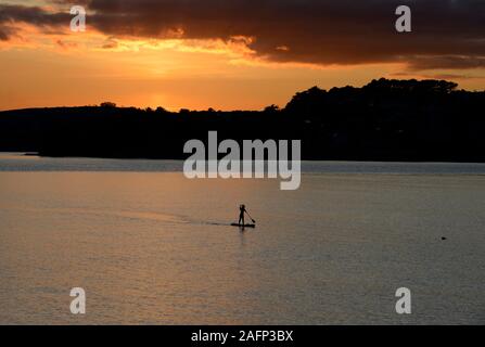 Un unico stand-up paddleboarder si avvicina al porto di Torquay in Devon in una giornata di mare calmo come il sole tramonta dietro di lei Foto Stock