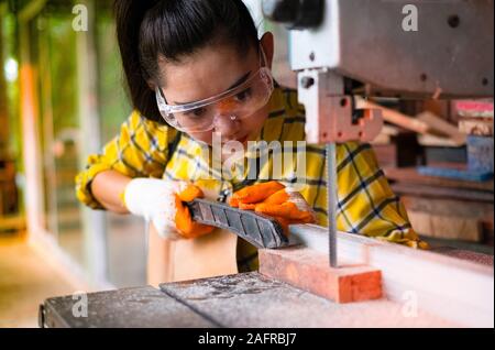 Le donne è permanente di lavoro artigianali legno tagliato in corrispondenza di un banco di lavoro con seghe a nastro power tools a carpenter la macchina in officina, lavoratore segare pannelli dalla wi Foto Stock