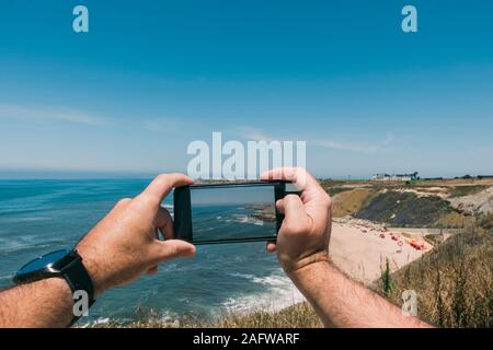 Personale prospettiva uomo con la fotocamera del telefono a fotografare soleggiato, scenic ocean Foto Stock