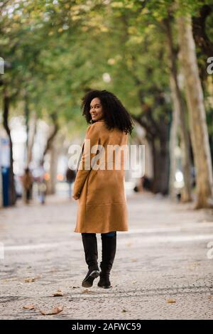 Ritratto fiducioso giovane donna guardando sopra la spalla sul marciapiede orlate da alberi Foto Stock