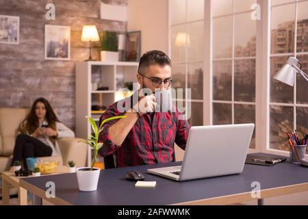 Barbuto imprenditore caucasico prendendo un sorso di caffè mentre si lavora sul computer portatile in salotto. Ragazza sul divano in background la navigazione sul telefono. Chip sul tavolo. Foto Stock