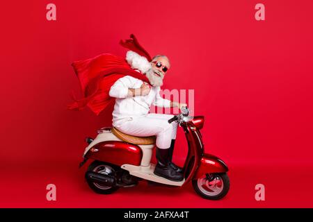 A piena lunghezza laterale del profilo fotografia del titolare di pensione o di rendita divertente borsa da trasporto con doni seduto sul motociclo fretta che indossa un maglione bianco stivali pantaloni isolato Foto Stock