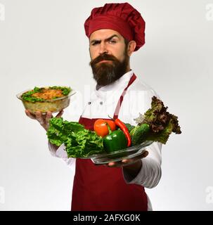 Alimentazione sana e concetto di cucina. Lo Chef detiene la coppa con Casseruola di patate e verdure. Uomo con la barba su sfondo bianco. Cucinare con un rigoroso volto in Borgogna mantiene uniforme il piatto cotto al forno e insalata mista Foto Stock