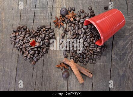 A forma di cuore ad gioiello in grani di caffè formando un cuore accanto a una fuoriuscita di rosso coppa piena di fagiolini sullo sfondo di legno Foto Stock