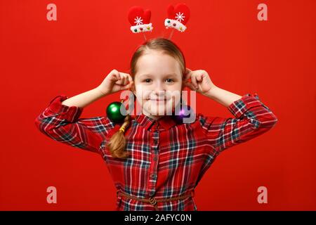 Funny felice bambina con orecchini palla di natale decorazione. Un bambino in un abito rosso e manopole con archetto su uno sfondo colorato. Foto Stock